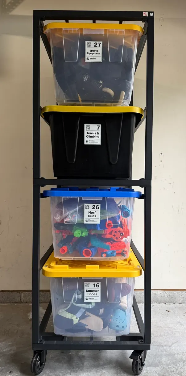A black rolling cart in a garage holding four storage bins, each with a Muninn label on its front: a QR code, a big number and a name. Top to bottom: 27 “Sports Equipment”, 7 “Tennis & Climbing”, 26 “Nerf Guns” and 16 “Summer Shoes”. Through the clear sides, sports gear, toy blasters and shoes.