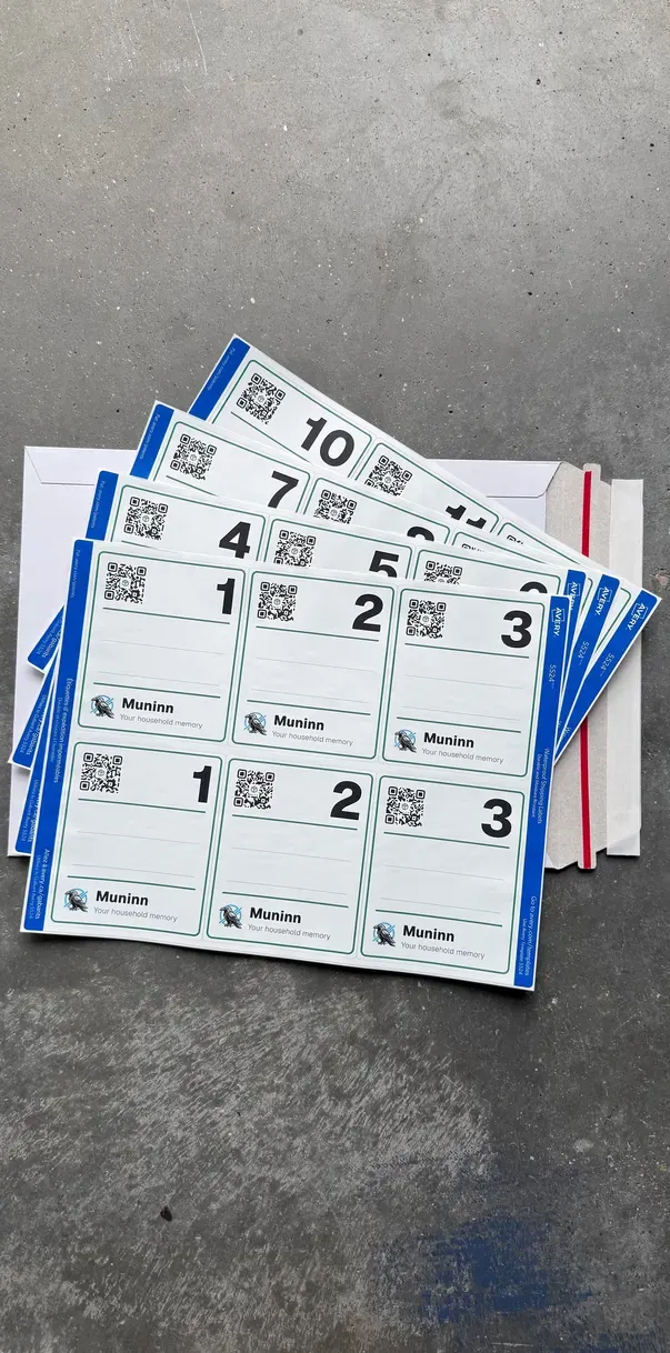A printed Muninn label pack fanned out on a concrete floor: four sheets of white labels with blue edges, numbered 1 to 12, two of each number. Every label has a QR code, a big bin number, blank lines to write on and the Muninn raven. The white mailer it came in is underneath.